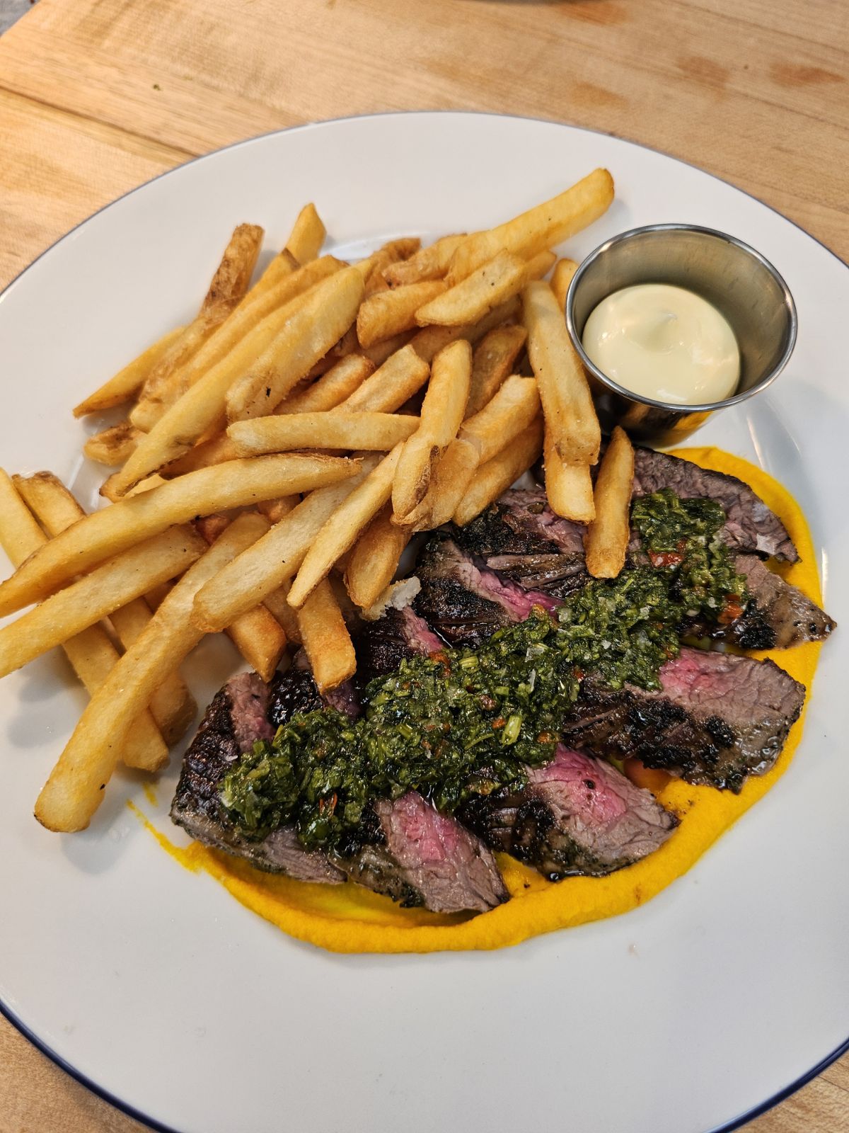 Images depicts neatly cut and arranged slices of flank steak topped with chimichurri and served with a side of frites.